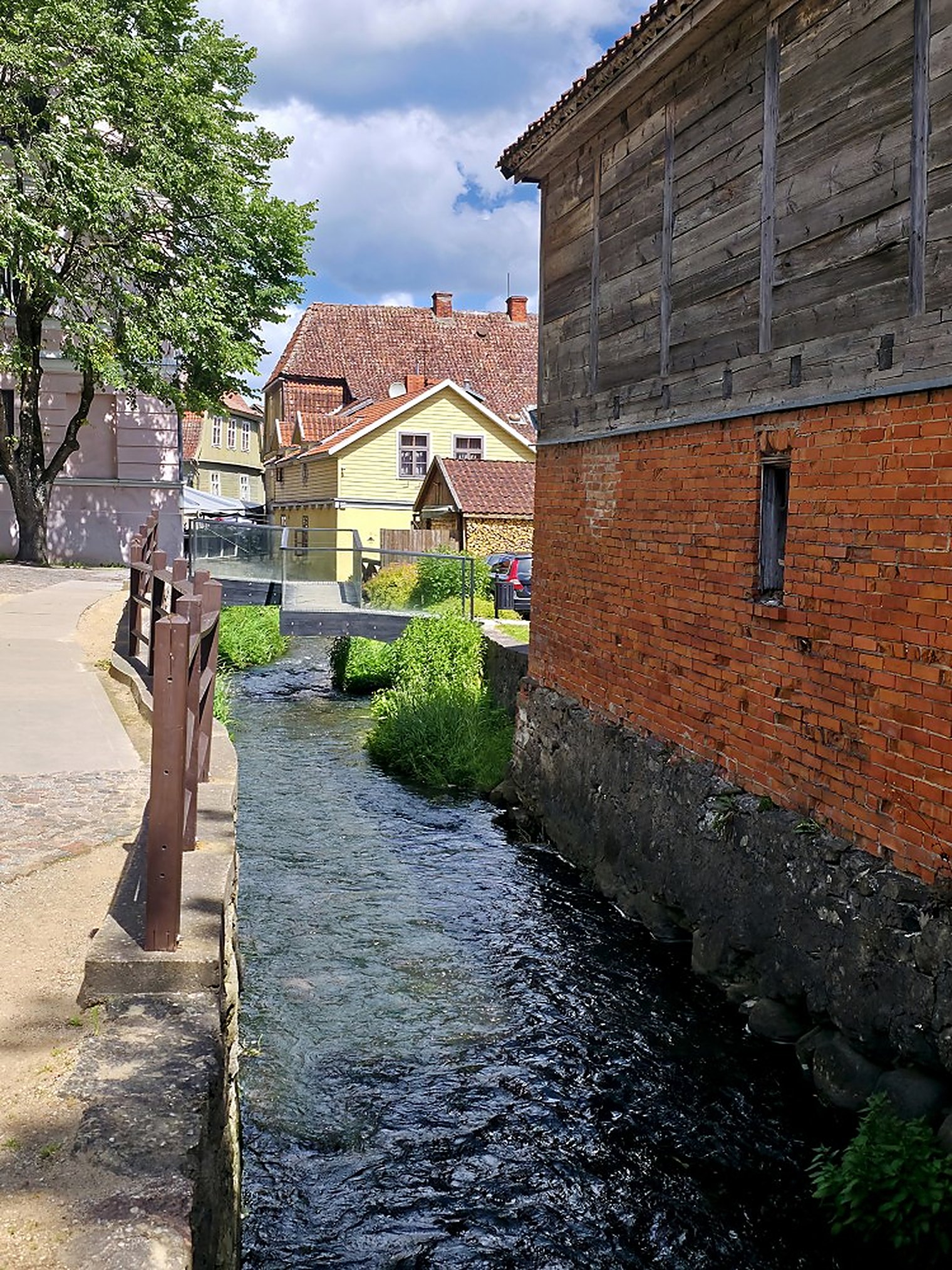 No bank, no railing. The Alekšupīte runs straight against the back walls, here beside the district council.